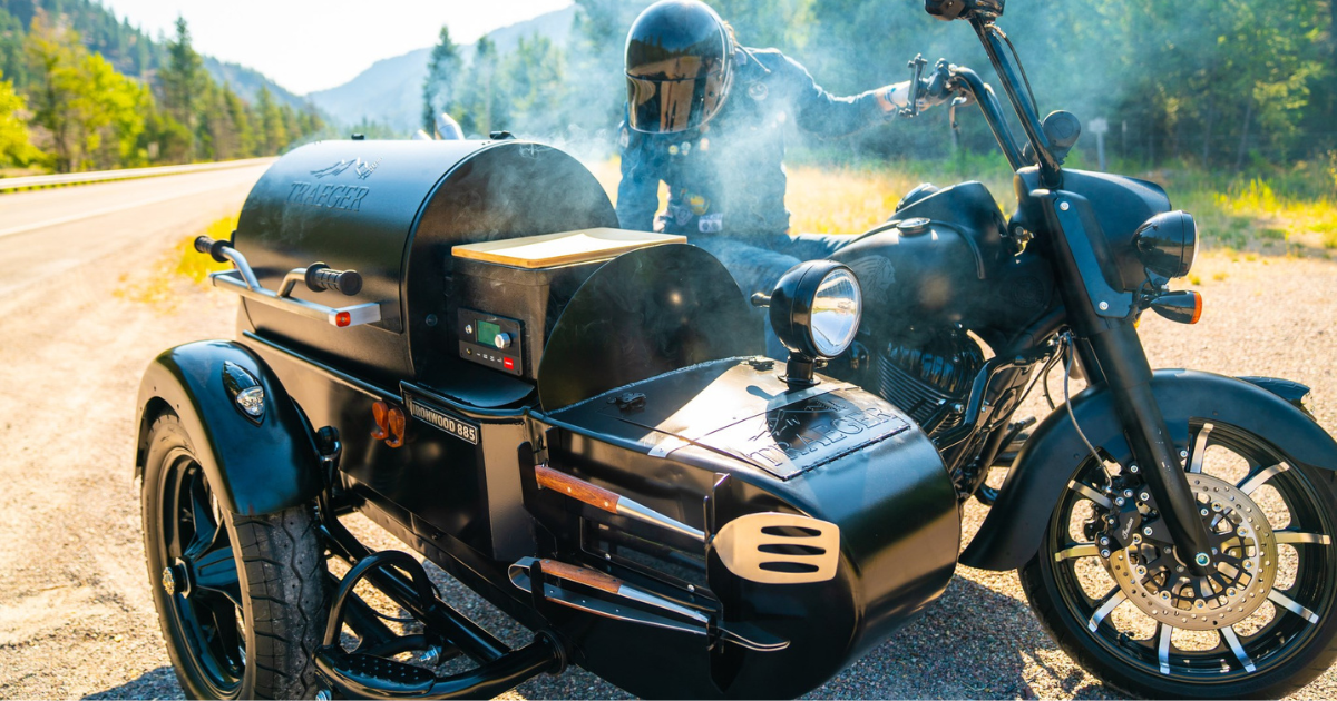 A matte black Indian motorcycle with a custom sidecar-mounted Traeger grill smokes gently by the roadside. A rider in full black gear leans on the bike, surrounded by pine trees and distant mountain peaks under a bright sky.